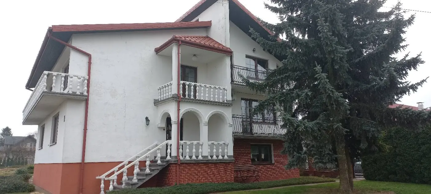 Piętrowy dom z białą elewacją, balkonami z białymi balustradami i ceglanym cokołem