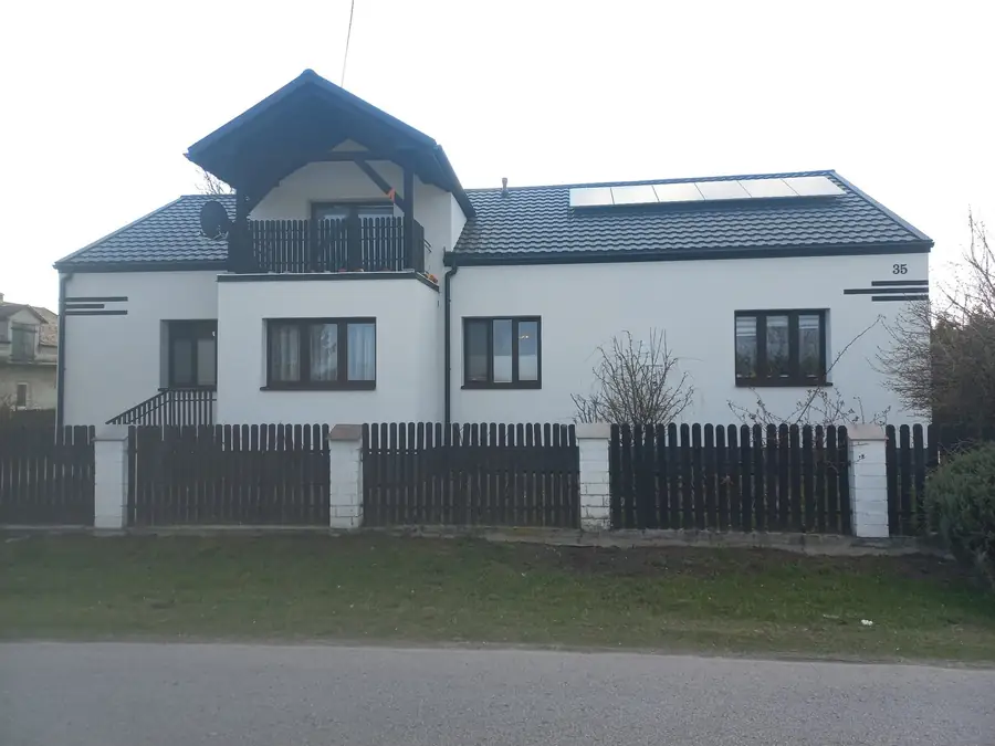 Długi dom z białą elewacją i balkonem w szczycie, przed nim drewniany płot